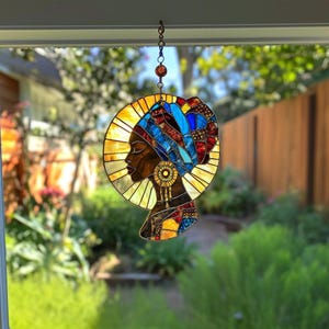 May include: Stained glass panel depicting a profile of a woman with dark skin wearing a blue and red head wrap and gold jewelry. The woman is set against a yellow and orange halo. The panel is suspended by a chain.