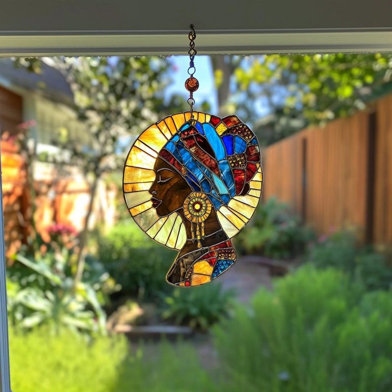 May include: Stained glass panel depicting a profile of a woman with dark skin wearing a blue and red head wrap and gold jewelry. The woman is set against a yellow and orange halo. The panel is suspended by a chain.