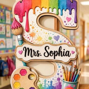 May include: A wooden hanging sign shaped like the letter S, decorated with rainbow paint drips, colorful splatters, and small hearts. It features a paintbrush, a paint palette with six colors, and a cup holding five brushes, with the text Mrs. Sophia.
