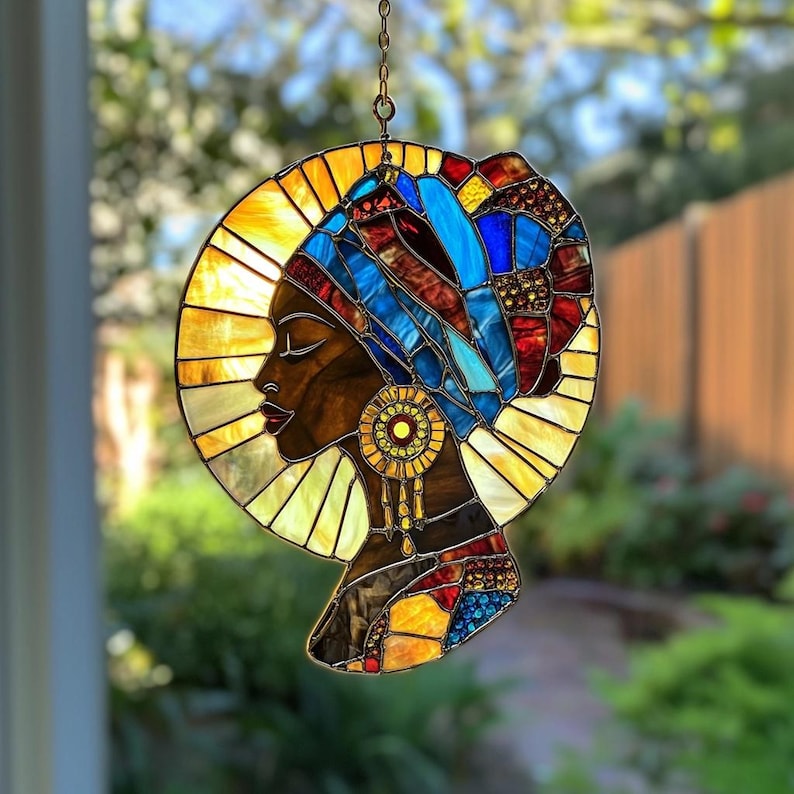 May include: Stained glass panel depicting a profile of a woman with dark skin, wearing a blue and red head wrap and gold earrings. The panel is set against a yellow and gold halo, with a chain for hanging.