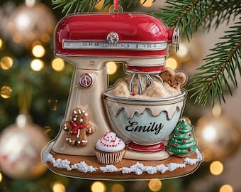 May include: A red and cream-colored stand mixer ornament featuring a mixing bowl with the name Emily printed on the front. The base is decorated with a gingerbread man, a cupcake, and a small Christmas tree, all accented with white snow.