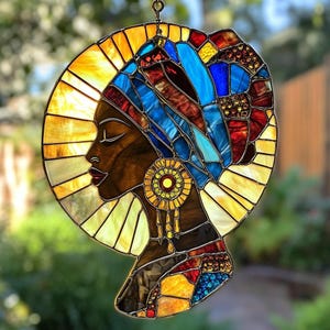 May include: Stained glass artwork depicting a profile of a woman with a colorful head wrap and earrings. The piece features a circular sunburst design in shades of yellow and orange, with a brown face and red lips. The head wrap is blue, red, and brown.
