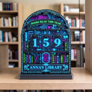 May include: A black and teal LED book counter with the text "BOOKS READ THIS YEAR" and "ANNA'S LIBRARY". The counter displays the number "159" in bright blue and pink. The design features a circuit board theme.
