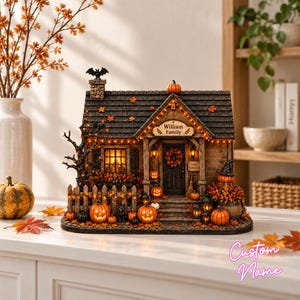 以下が含まれることがあります： ハロウィーンをテーマにした装飾的なミニチュアハウス。暗い色の屋根、William Familyと書かれた看板、オレンジ色のイルミネーションライト、多数の彫刻されたカボチャ、黒猫、煙突の上のコウモリ、そして秋の紅葉が特徴です。