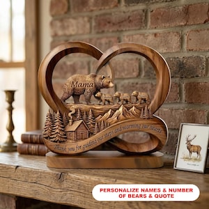 Op de afbeelding: Een houten sculptuur met een berenfamilie, met het woord "Mama" en de namen "Emma", "Liam" en "Noah". De sculptuur bevat een hut, bomen en bergen, met de tekst "Thank you for guiding our steps and warming our hearts".