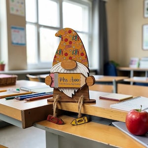 Puede incluir: Decoración de gnomo de madera con un sombrero estampado con manzanas, libros y estrellas. El gnomo sostiene un lápiz con el texto "Mrs. Anne" y "Tiny Humans, Big Hearts". Dos adornos de crayones cuelgan debajo. El gnomo está en un escritorio.