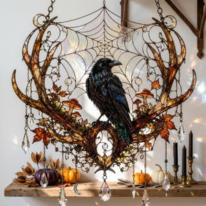 May include: A decorative wall hanging featuring a central black raven perched on brown antlers. The piece includes a wire spiderweb, iridescent accents, orange autumn leaves, small mushrooms, and hanging clear crystal prisms on metal chains.