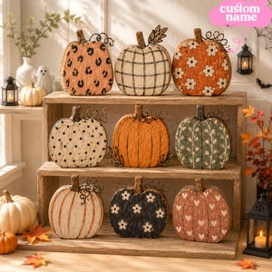 Può includere: Una collezione di nove zucche decorative in legno disposte su uno scaffale in legno a tre livelli. Ogni zucca presenta un motivo unico, tra cui stampa leopardata, quadri, fiori, pois, arancione tinta unita, viticci, strisce e cuori.