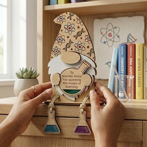 Puede incluir: Regalo de agradecimiento para profesores con forma de gnomo de madera. El gnomo tiene un sombrero estampado con diseños de temática científica, una barba blanca y un letrero en forma de corazón que dice "To Teacher Anne: For sparking the magic of learning." El gnomo tiene dos piernas en forma de vaso de precipitados.