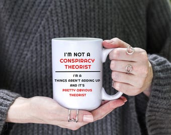 May include: White ceramic coffee mug with black text that reads "I'm not a conspiracy theorist I'm a things aren't adding up and it's pretty obvious theorist".