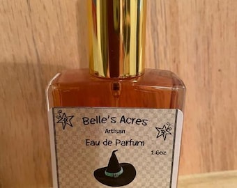 May include: A rectangular glass perfume bottle filled with amber liquid, topped with a shiny gold cap. The label features the text Belle's Acres, Artisan, Eau de Parfum, 1.6oz, and Witches Brew, with a graphic of a black witch hat and stars.