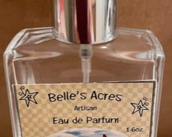 May include: A clear glass bottle of Mountain Woman perfume by Belle's Acres. The bottle is labeled with a brown and white label with a mountain scene and the text "Belle's Acres Artisan Eau de Parfum 1.6oz Mountain Woman".