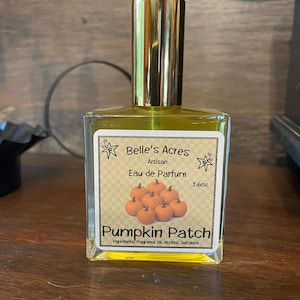 Puede incluir: Un frasco de perfume de vidrio rectangular con tapa dorada y líquido amarillo. La etiqueta dice Belle's Acres Artisan Eau de Parfum 1.6oz Pumpkin Patch con una ilustración de calabazas pequeñas y una lista de ingredientes.