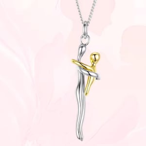 Op de afbeelding: Zilverkleurige hangerketting met een gestileerd omarmingsontwerp. De hanger toont twee figuren, één zilver en één goud, in een omhelzing. De ketting heeft een delicate ketting en staat tegen een zachte roze achtergrond.
