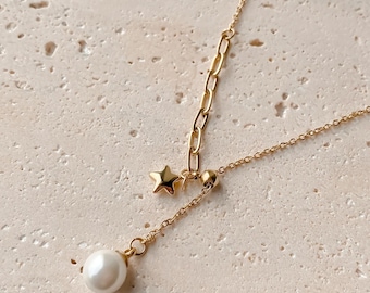 May include: A gold-colored necklace with a delicate chain design. The necklace features a small star charm and a dangling white pearl pendant. The chain has a unique link pattern near the star. The background is a light beige color.