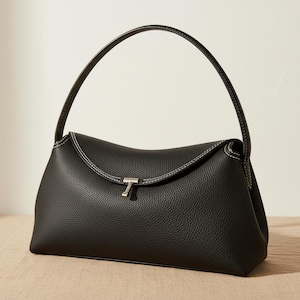 May include: Black handbag with a curved top and a single handle. The bag features white stitching along the edges and a silver-toned clasp. The leather-like material has a textured appearance, and the bag is set against a neutral background.