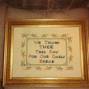 May include: Framed cross-stitch artwork with gold-colored frame. The text reads "WE THANK THEE THIS DAY FOR OUR DAILY BREAD" in black and red thread. The artwork has a cream-colored background with decorative border.