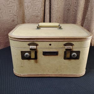 May include: A vintage beige suitcase with a tan handle and trim. The rectangular case has two metal latches with keyholes and a small nameplate. The suitcase is closed and appears to be made of a durable material.