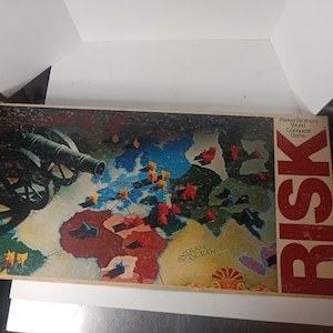 May include: Vintage Risk board game by Parker Brothers. The game board depicts a world map with territories marked by small plastic pieces. The box features a cannon illustration and the game's title in bold letters.