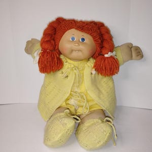 以下が含まれることがあります： 赤い三つ編みと青い目のヴィンテージキャベツパッチキッズ人形。人形は、セーター、ロンパース、ブーティーを含む黄色の衣装を着ています。人形の腕は伸ばされています。