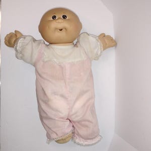 Könnte beinhalten: Eine Vintage-Cabbage-Patch-Kid-Puppe mit kahlem Kopf, bemalten Gesichtszügen und einem rosa-weiß karierten Strampler mit Spitzenbesatz. Die Arme der Puppe sind ausgestreckt, und sie hat einen neutralen Gesichtsausdruck.
