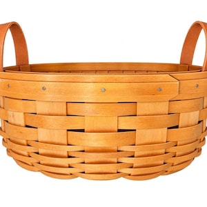 May include: A round woven wooden basket featuring a light tan finish, a wide reinforced rim secured with small metal rivets, and two sturdy upright handles made of a matching tan material attached to the sides of the basket structure.