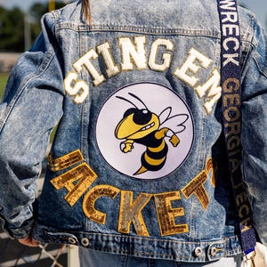 Może przedstawiać: Jasnoniebieska kurtka dżinsowa z napisem "STING EM JACKET" złotymi i białymi literami. Duża okrągła naszywka przedstawia pszczołę z kreskówki. Widoczny jest pasek białej torebki.