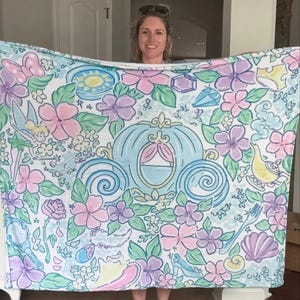 May include: A white blanket featuring a whimsical design with a light blue Cinderella carriage, pink and purple flowers, and other fairytale-themed illustrations. The blanket is held up, showcasing its full pattern.