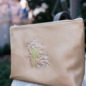 Könnte beinhalten: Eine goldfarbene Reißverschlusstasche mit einem gestickten Design einer grünen Fee und weißen Sternen. Die Tasche besteht aus einem strukturierten Material und liegt auf einer Steinoberfläche.