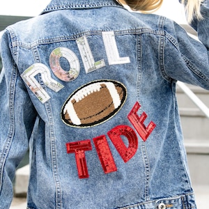 May include: A light-wash denim jacket with the words "ROLL TIDE" appliqued on the back. The letters are embellished with sequins. A football-shaped patch is centered between the words. The jacket has a button-down front and pockets.