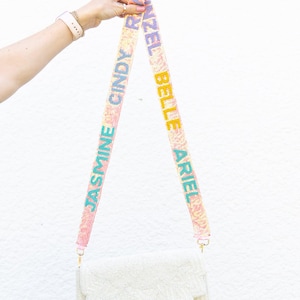 Puede incluir: Un bolso de mano blanco con cuentas y solapa floral, sostenido por una correa con los nombres "JASMINE", "CINDY", "ARIEL" y "BELLE" en colores vibrantes. El bolso tiene herrajes dorados y un diseño decorativo.