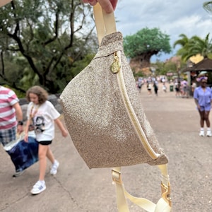 May include: A gold glitter fanny pack with a white strap and a gold zipper. The fanny pack has a small gold heart charm with the letters "DL" on it.