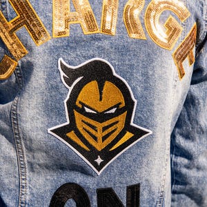 Puede incluir: Chaqueta vaquera de lavado claro con letras doradas con lentejuelas que deletrean "CHARGE" en la espalda. Un parche con el emblema de un caballero negro y dorado está centrado debajo de las letras, con la palabra "ON" en lentejuelas negras debajo.