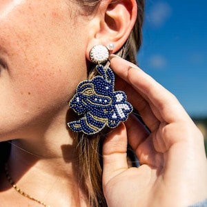 May include: Navy blue beaded earrings with a floral design. The earrings have white and gold accents, and a white beaded stud. A gold necklace with a pearl pendant is also visible.
