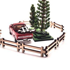 May include: A miniature red Chevrolet pickup truck with a white tailgate, parked near two model evergreen trees. The scene is enclosed by a small wooden fence, creating a diorama effect. The truck is the focal point of this miniature landscape.