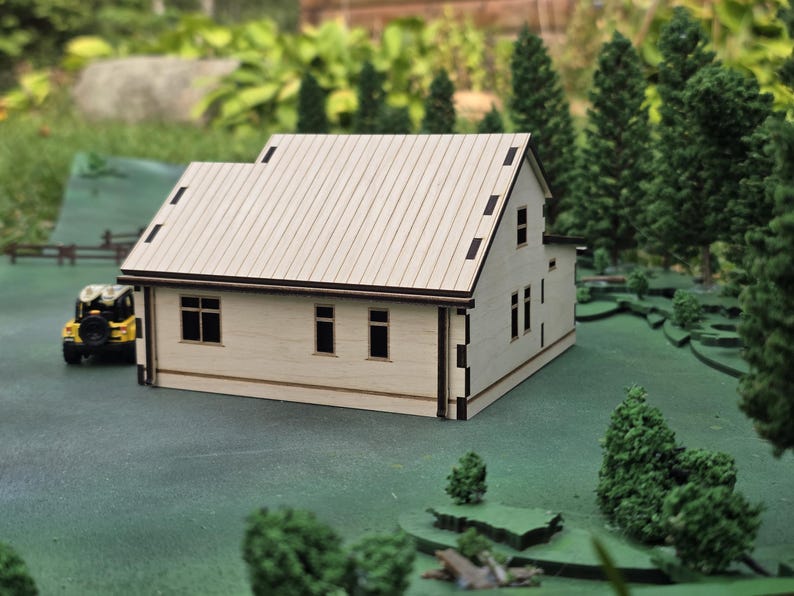 May include: A miniature wooden house with a light-colored exterior and a contrasting dark brown roof and trim. The model has multiple windows and is set on a green surface with miniature trees and a yellow toy vehicle. The house is a detailed architectural model.