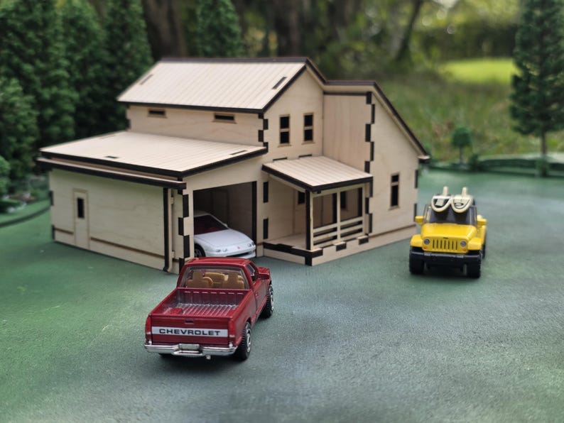 May include: A miniature wooden house with a garage, a red Chevrolet pickup truck, a white car in the garage, and a yellow Jeep toy. The house has a light wood finish with black trim. The scene is set on a green surface.