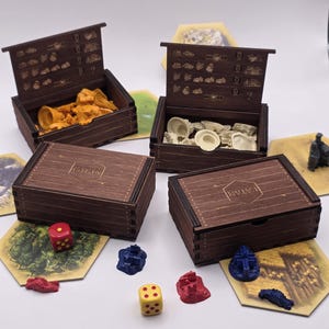 May include: Two dark brown wooden storage boxes with the Catan logo engraved on the lids. One box is open, revealing yellow game pieces, while the other is open, showing white pieces. Two closed boxes, red and yellow dice, and game tiles are included.