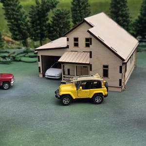 May include: A miniature wooden house with a garage, a yellow toy Jeep with a surfboard on top, and a red toy car. The house has a light-colored roof and walls, with dark trim. The scene is set on a green surface with miniature trees in the background.