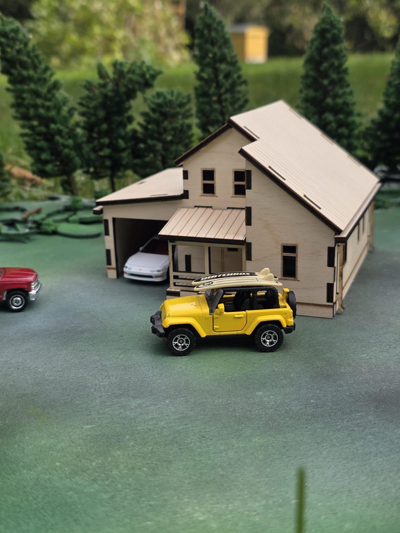 May include: A miniature wooden house with a garage, a yellow toy Jeep with a surfboard on top, and a red toy car. The house has a light-colored roof and walls, with dark trim. The scene is set on a green surface with miniature trees in the background.