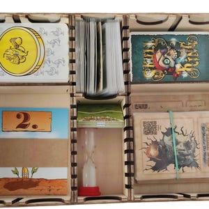 Puede incluir: Un organizador de juegos de mesa de madera con múltiples compartimentos que contienen varios componentes de juego, incluyendo mazos de cartas con una moneda de oro y una calavera, una carta marcada con un 2, un pequeño reloj de arena y un mazo Spellfire Re-Master The Magic.