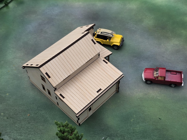 May include: A miniature wooden house with a light brown finish, featuring a sloped roof and rectangular windows. A yellow toy SUV with a surfboard and a red pickup truck are nearby. The scene is set on a green surface.