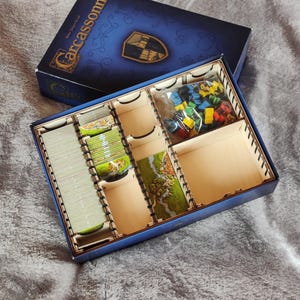 May include: A Carcassonne board game box with a blue lid and an open interior featuring a wooden organizer insert. The insert contains stacks of square cardboard tiles with landscape illustrations and a plastic bag filled with small wooden game pieces.