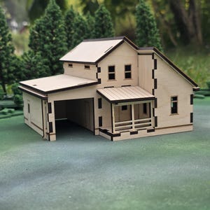 May include: A miniature wooden house model with a garage, porch, and multiple windows. The house is constructed from light-colored wood with dark brown trim, and sits on a green surface. The model has a two-story design with a gabled roof.