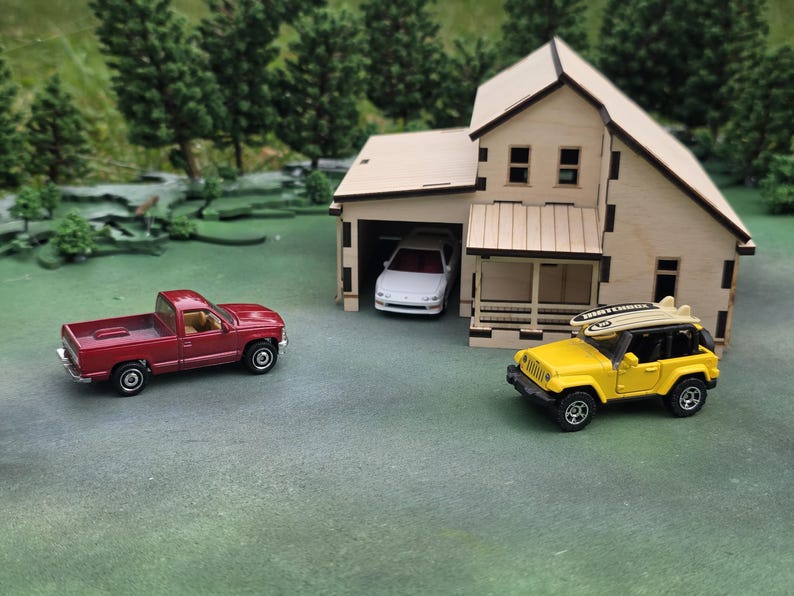 May include: A miniature scene featuring a red pickup truck, a yellow Jeep with a surfboard, and a wooden house with a garage. A white car is parked inside the garage. The scene is set on a green surface with miniature trees.