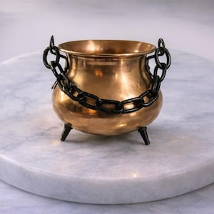 May include: A small, shiny, copper-coloured cauldron with a black chain handle and three black legs. The cauldron is sitting on a white marble-like surface. The cauldron is a decorative item.