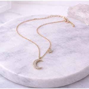 May include: A gold necklace with a crescent moon pendant and a star charm, both set with small, clear gemstones. The necklace is displayed on a white marble surface, with a clear crystal in the background.