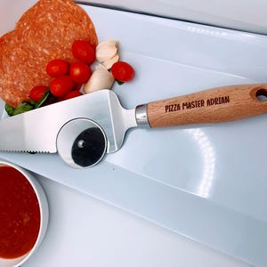 May include: A white ceramic plate with a pizza cutter, salami, cherry tomatoes, garlic cloves, and a small bowl of red sauce. The pizza cutter has a wooden handle with the text "Pizza Master Adrian" engraved on it.