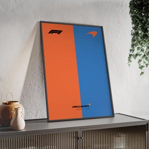 Peut inclure: Affiche encadrée avec un design divisé verticalement. Un côté est orange, l'autre est bleu. Les logos et le texte noirs incluent le logo F1, un logo McLaren et "MCLAREN FORMULA 1 TEAM". L'affiche est affichée sur un meuble gris.