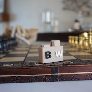 Op de afbeelding: Een beige houten schaakbord met zwarte en gouden schaakstukken. Een klein beige blokje met de letters "B" en "W" in zwart en wit staat op het bord.
